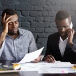 Candid shot of serious Afro-American colleagues in formal wear working together in office: man in shirt looking through papers while male wearing glasses having phone conversation, looking worried